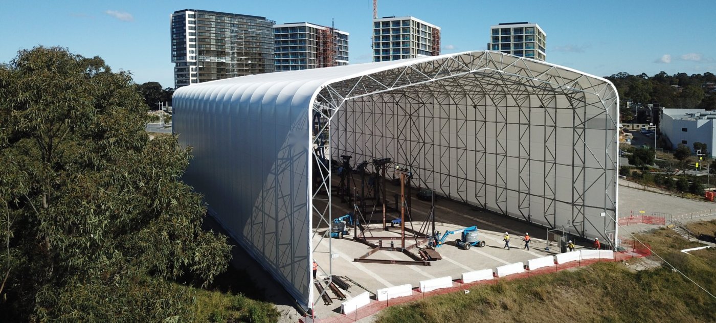 Large free standing steel and fabric structure on site with workers and lifts inside, beside apartment towers